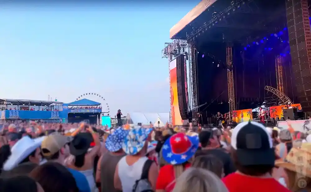 Crowd at an outdoor concert with a large stage and Ferris wheel in the background.