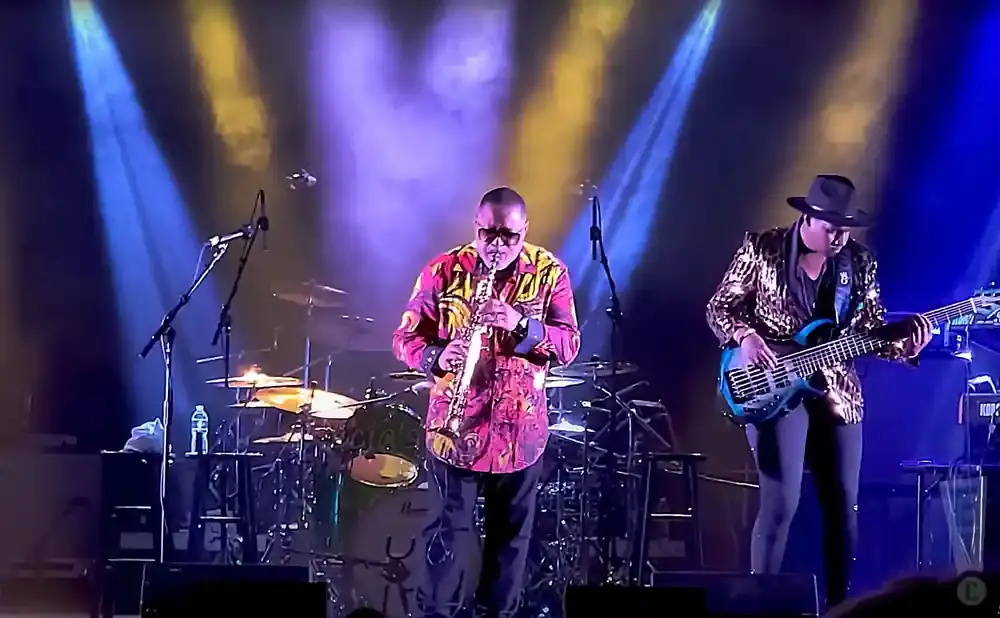 Musicians perform on a stage with colorful lights. A saxophonist in a vibrant shirt stands center, while a bassist in a sequined jacket plays to his right.