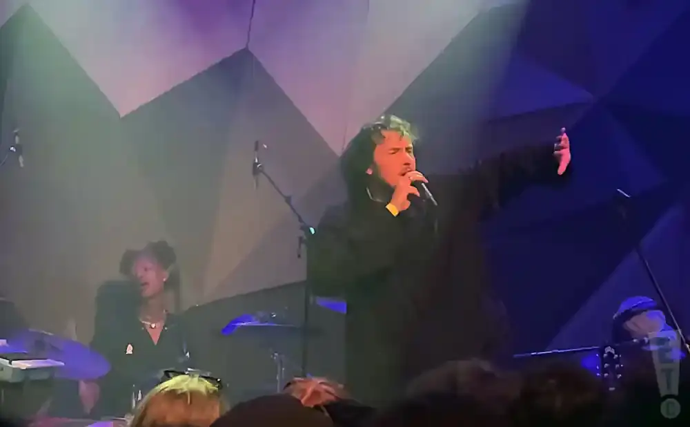 Singer with dark curly hair holds microphone and gestures with right hand, while a drummer plays on the left.