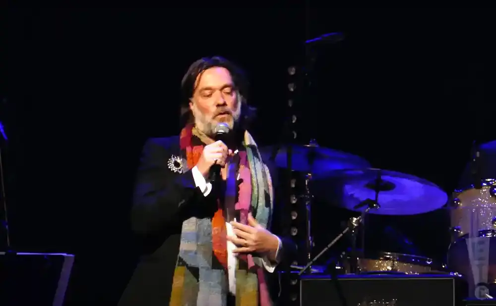 A man with a beard and scarf sings into a microphone on stage, with drums in the background.