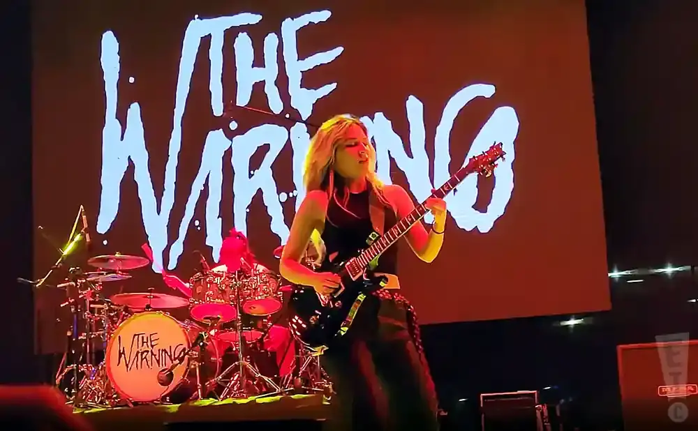 A female guitarist plays a black electric guitar in front of a large screen displaying 