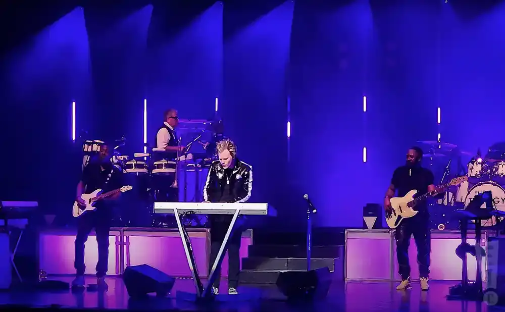 Musicians on stage playing instruments under blue lights, including a keyboardist, guitarist, and drummer.