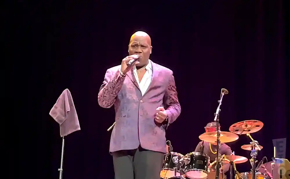 Singer in a purple jacket with a patterned lapel sings into a microphone on stage with a drummer in the background.
