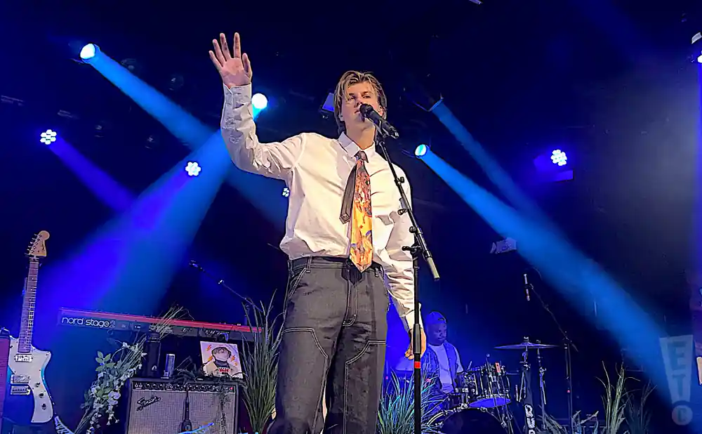 A male singer in a white shirt and tie waves to the audience during a blue-lit concert.