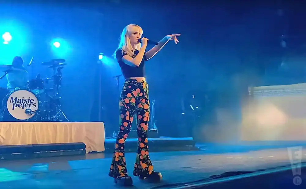 A singer with blonde hair wears a black top and floral bell-bottom pants, singing into a microphone on a blue-lit stage.