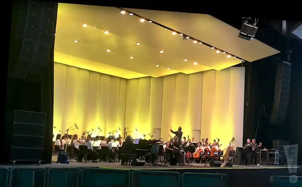 An orchestra performs on a brightly lit stage with a conductor directing.