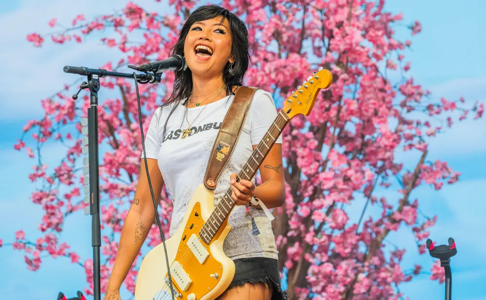 A woman joyfully sings into a microphone while playing a guitar on stage with a pink floral backdrop.