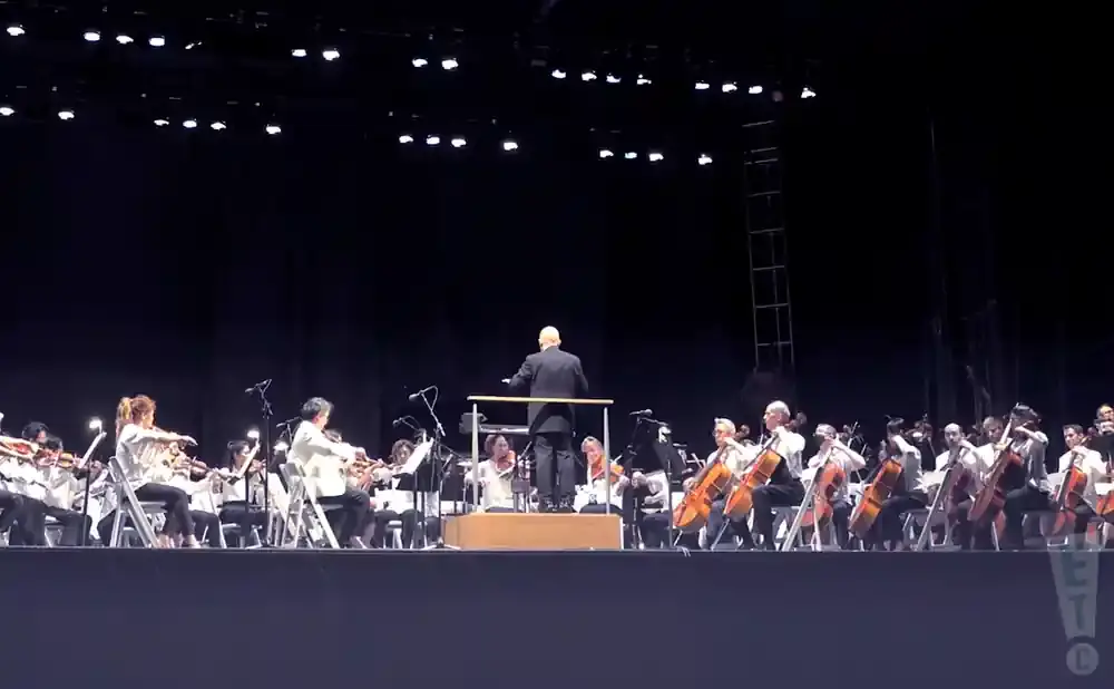 An orchestra performs on a stage, with a conductor leading them.