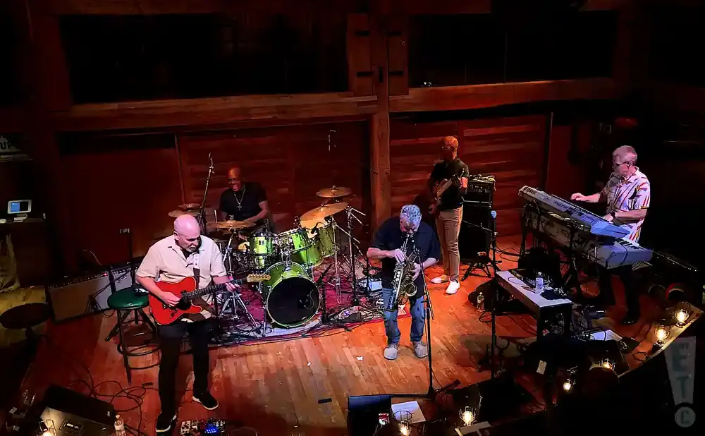 Band performing live music on stage, with a guitarist, drummer, saxophonist, and keyboardist.