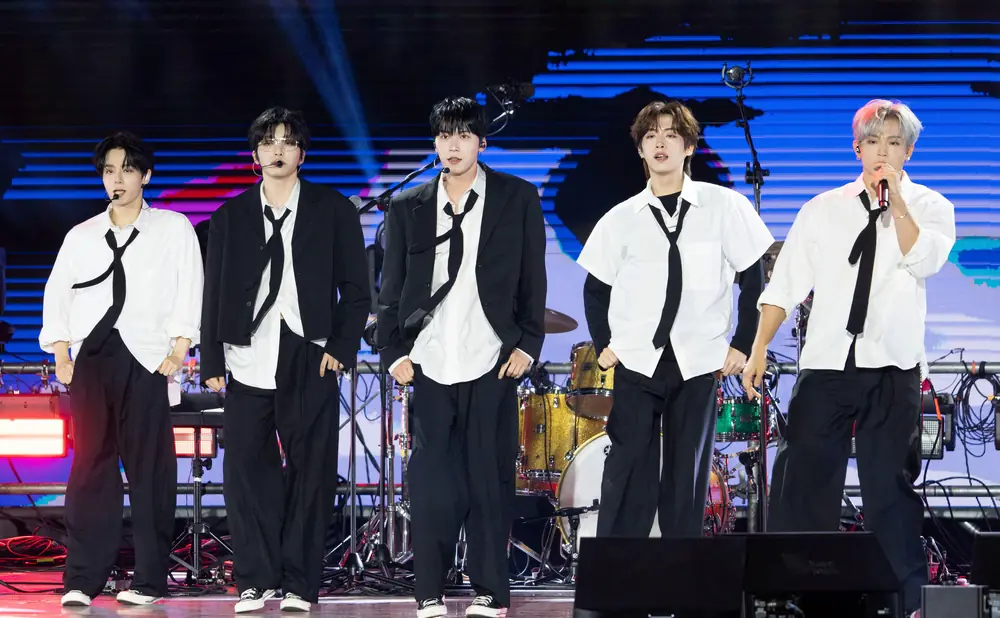 Five young men in black suits and white shirts with black ties perform on stage.