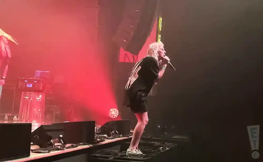 A female singer with blonde hair performs on stage with red lighting and speakers.