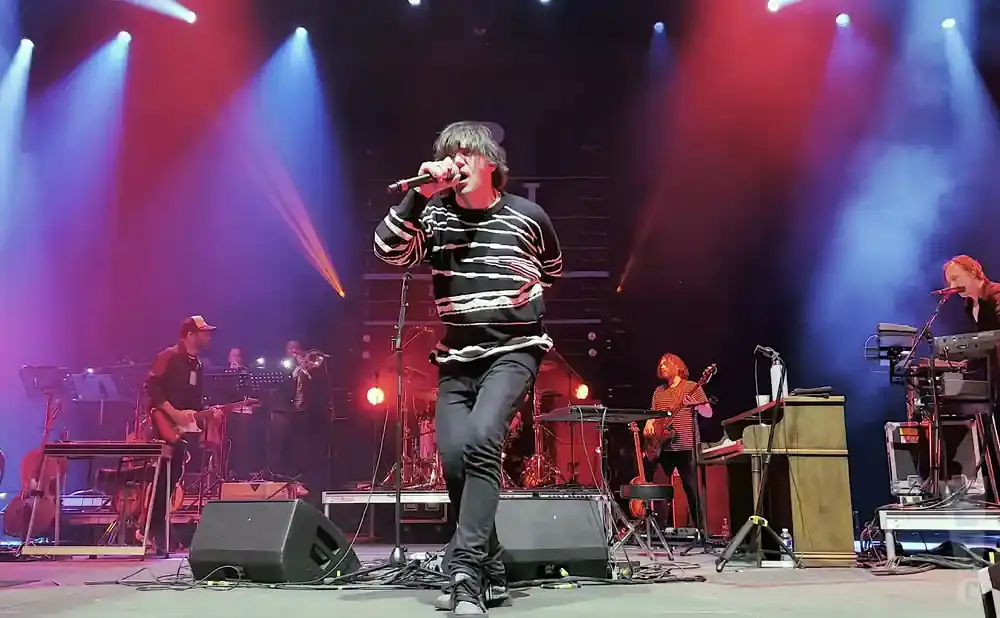 A band performs on stage with blue and red lights, a singer in a striped sweater in the foreground.