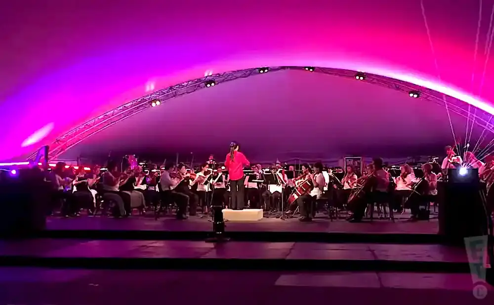 An orchestra performs under a large, pink, arched tent, illuminated by purple lights.