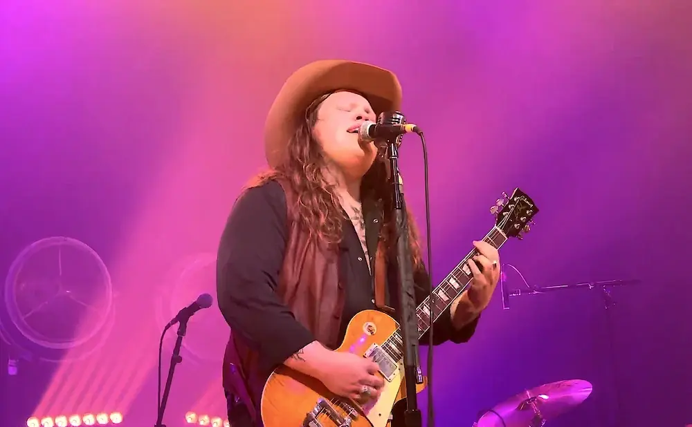 A person in a cowboy hat sings and plays a guitar on a stage lit by purple lights.