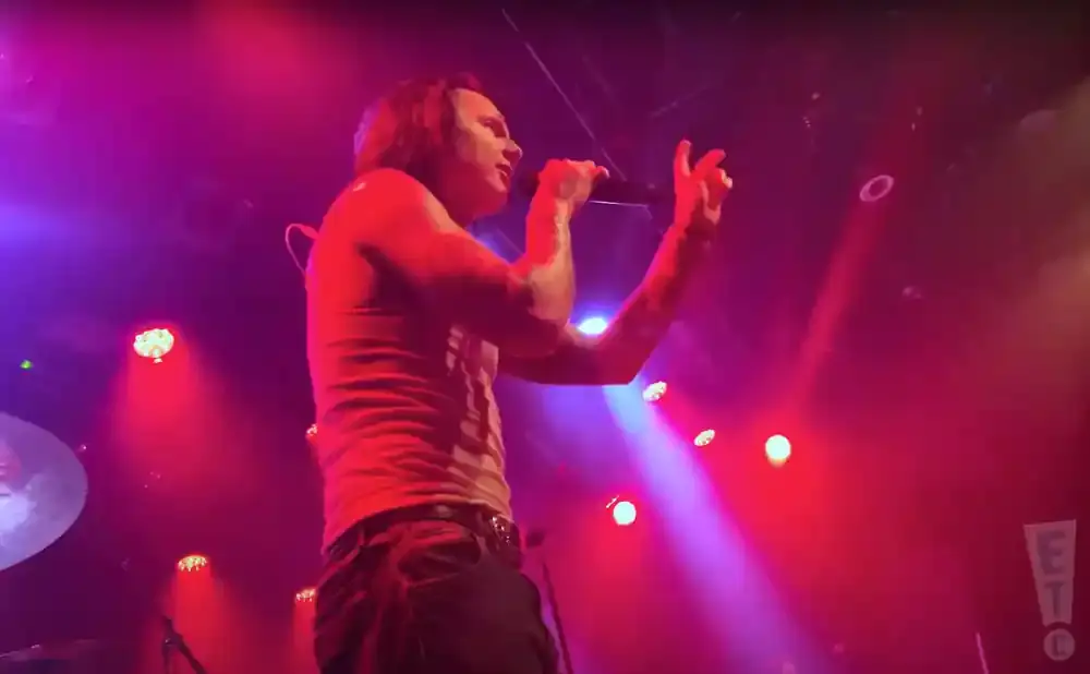 A male singer with long hair performs on a dimly lit stage, holding a microphone and gesturing with his hands, surrounded by red and purple stage lights.