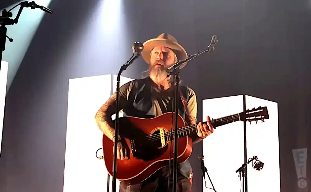 Musician plays an acoustic guitar on stage with a hat and beard.