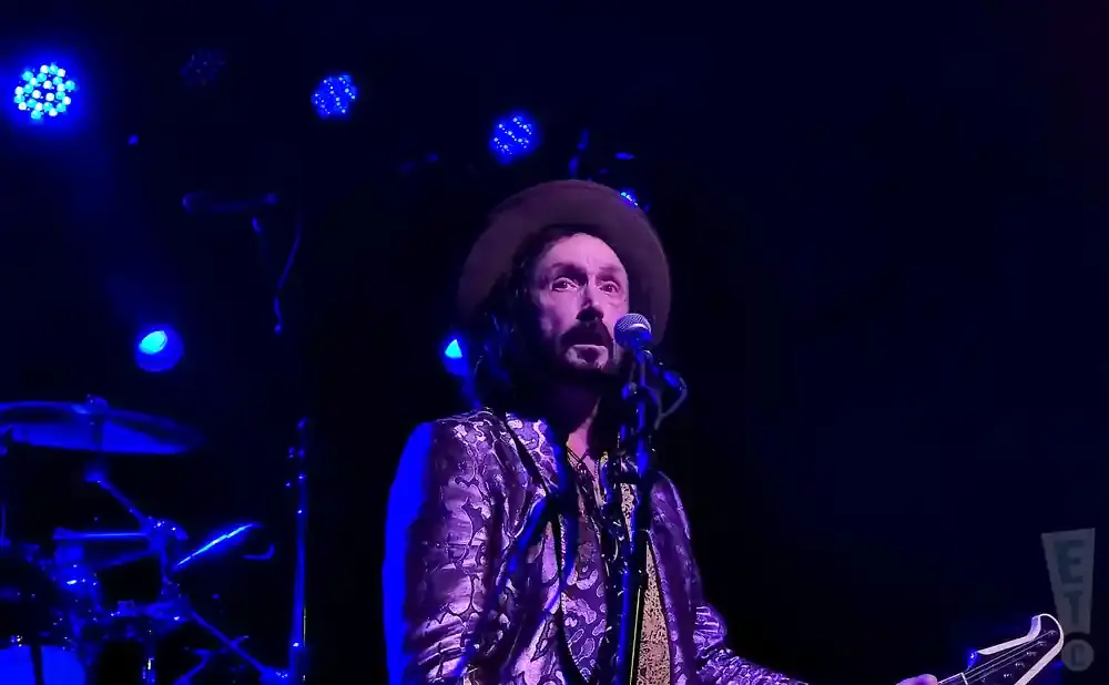 Man with hat and patterned jacket sings into microphone on a dark stage with blue lights.