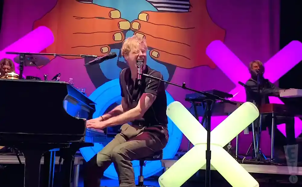 A male pianist sings and plays a grand piano on stage with a colorful backdrop.
