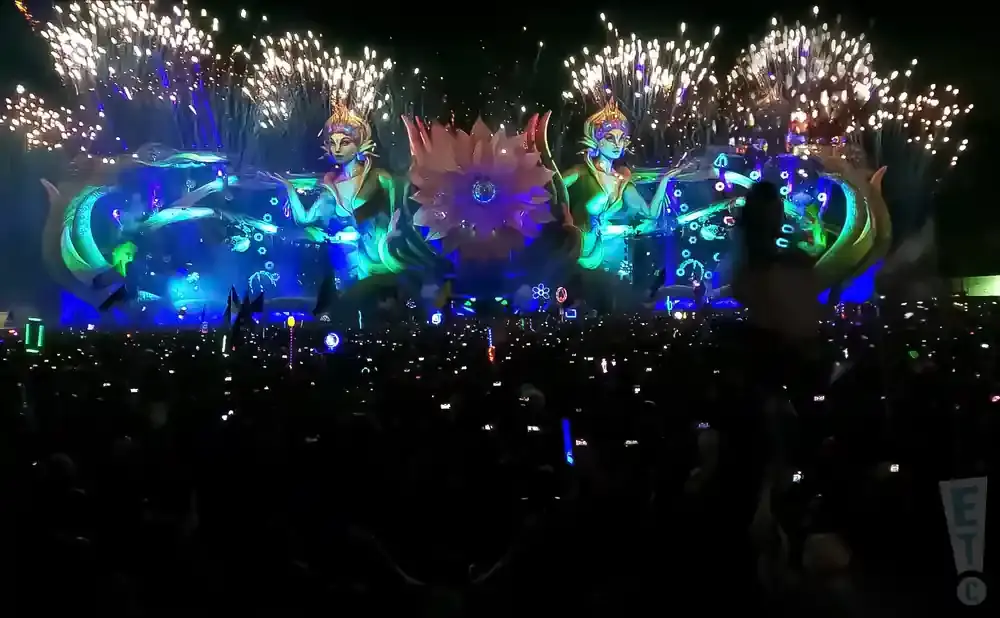 Fireworks erupt over a large crowd at a nighttime festival, with a fantastical stage featuring mermaid-like figures and a giant flower.