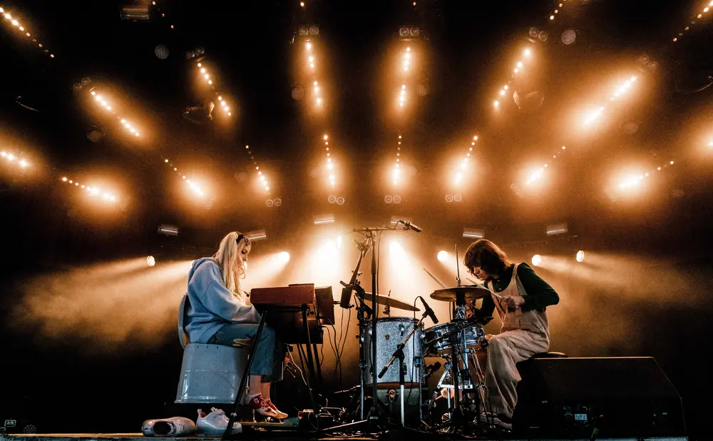 Two musicians perform on a stage lit by warm spotlights. A woman plays a keyboard on the left, and a person plays drums on the right.