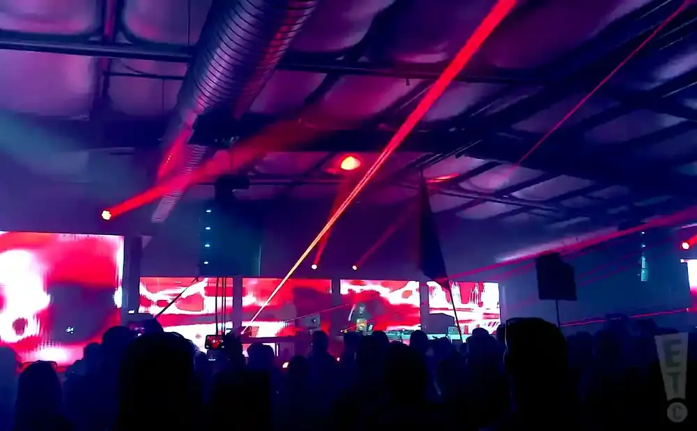 A crowd watches a DJ perform with red laser lights and a large screen displaying abstract red patterns.