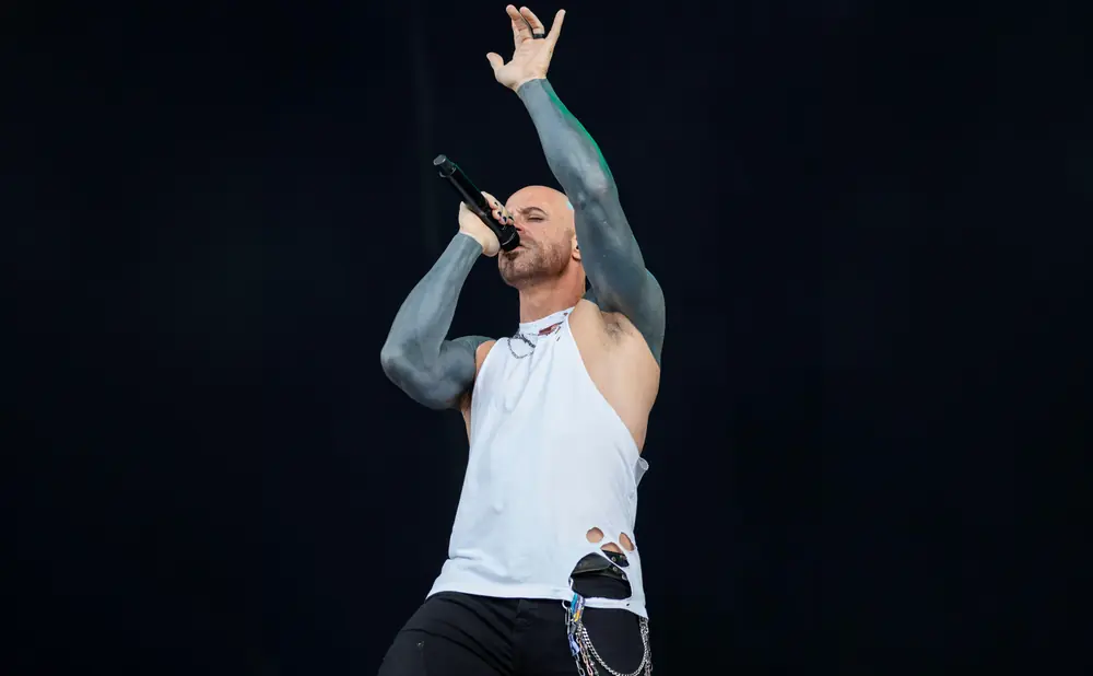 A bald man with tattooed arms sings into a microphone on stage, wearing a torn white tank top and black pants.