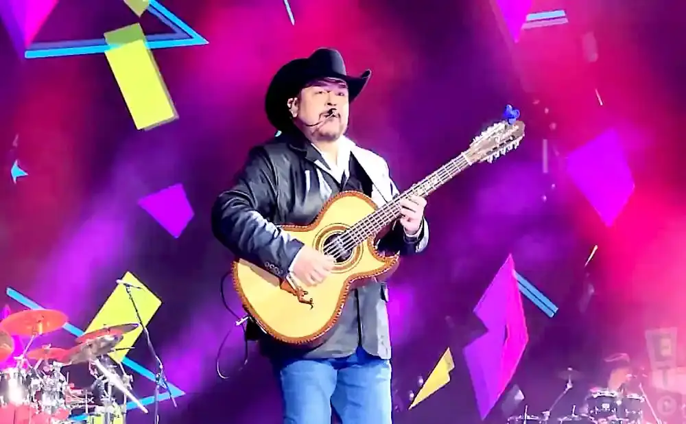 A man in a cowboy hat plays a guitar on a stage with purple and pink lighting and geometric shapes.