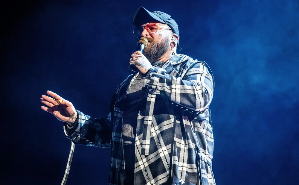 A man in a plaid jacket and cap sings into a microphone on a dark stage.