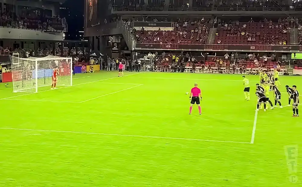 A soccer referee in pink stands near a ball, with players in black and yellow lined up for a free kick in front of a stadium crowd.