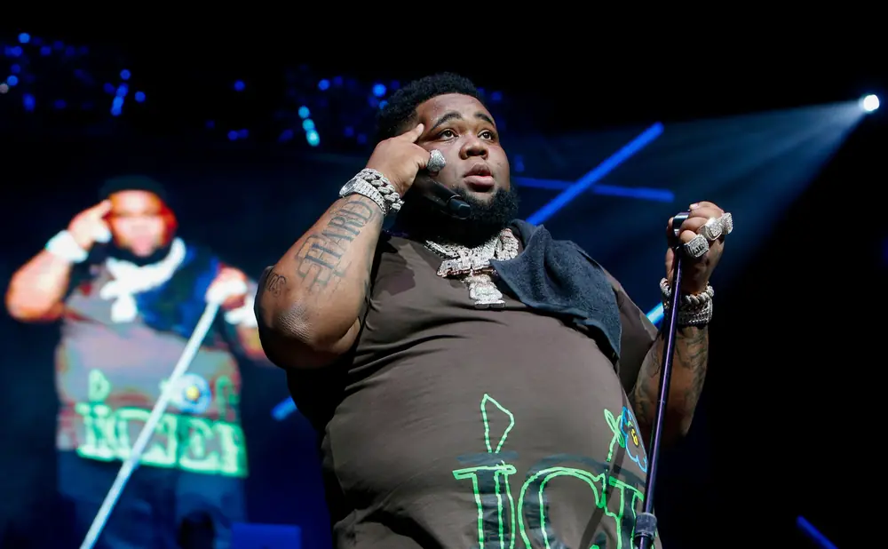 Rapper Rod Wave performs on stage wearing a brown t-shirt with green lettering, large diamond chains, and rings.