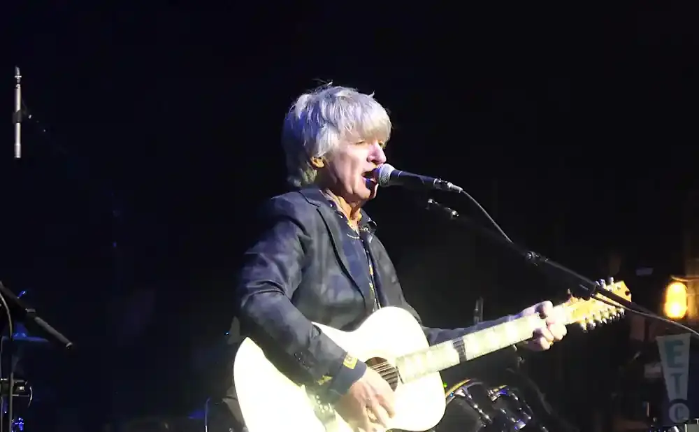 Musician with grey hair playing a white acoustic guitar and singing into a microphone on stage.