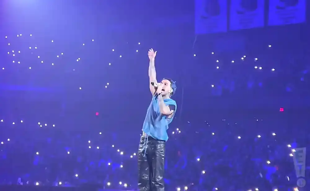 A male singer with blue hair sings into a microphone on a stage, with an audience holding up lights in the background.