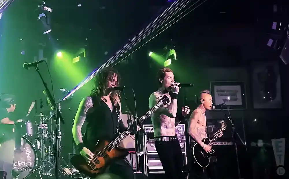 Three male musicians perform on a dimly lit stage with green and purple lights. One sings into a microphone, another plays bass, and the third plays gui…