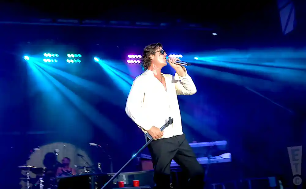 A man in a white shirt sings into a microphone on a stage with blue spotlights.