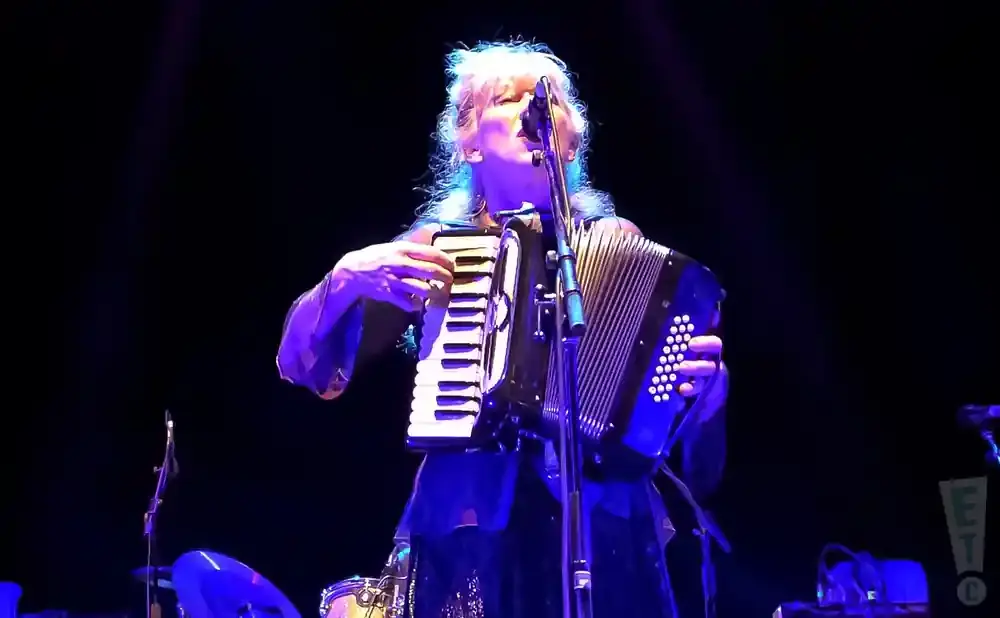 A person with blonde hair plays an accordion on a dark stage, bathed in purple light.
