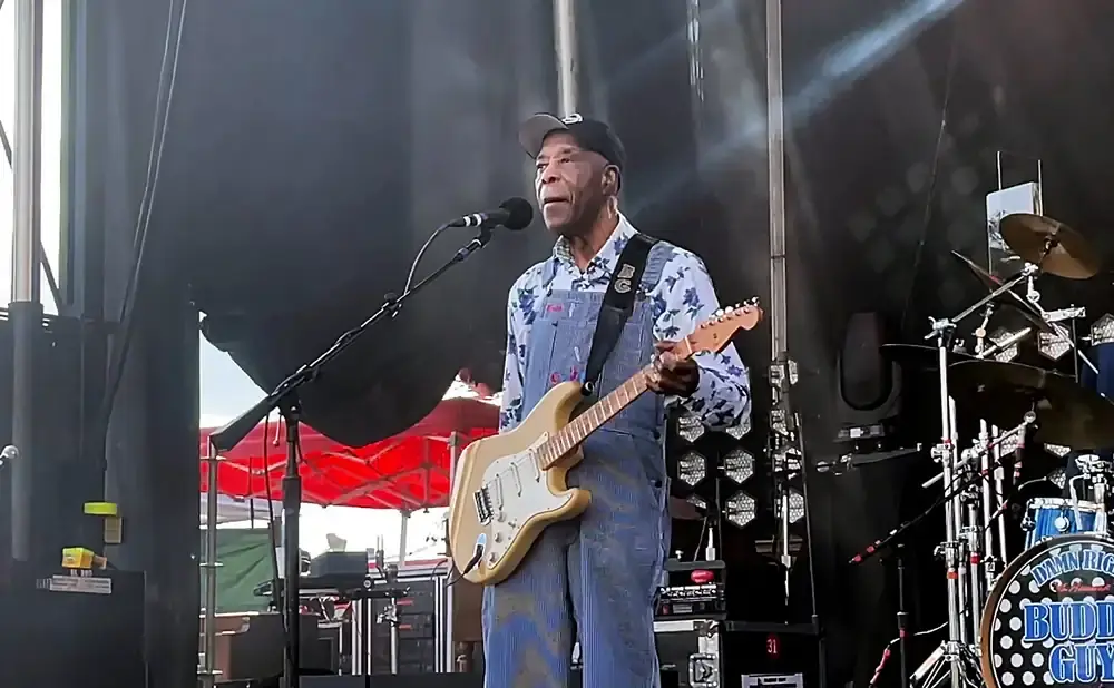 Buddy Guy performs on stage wearing a floral shirt and overalls, playing a light-colored electric guitar.