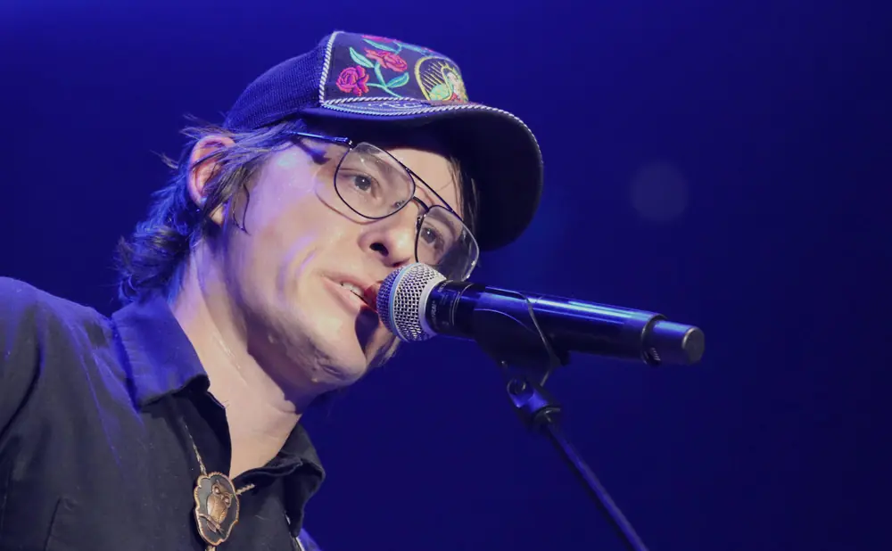 A man in a black shirt and hat sings into a microphone on a blue-lit stage.