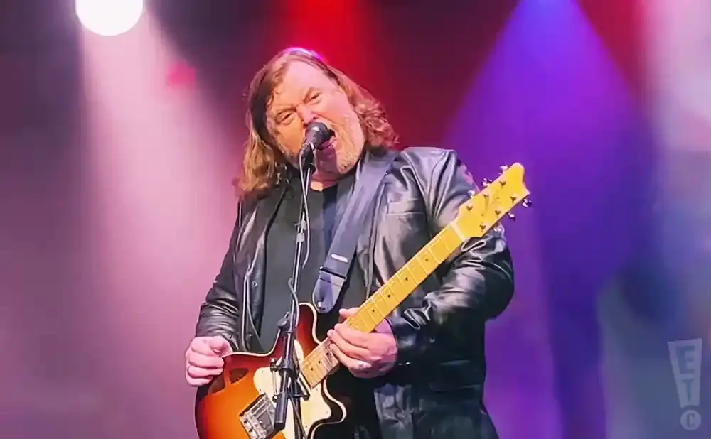 A man with long brown hair and a beard plays a sunburst guitar and sings into a microphone on a stage with purple and pink lights.