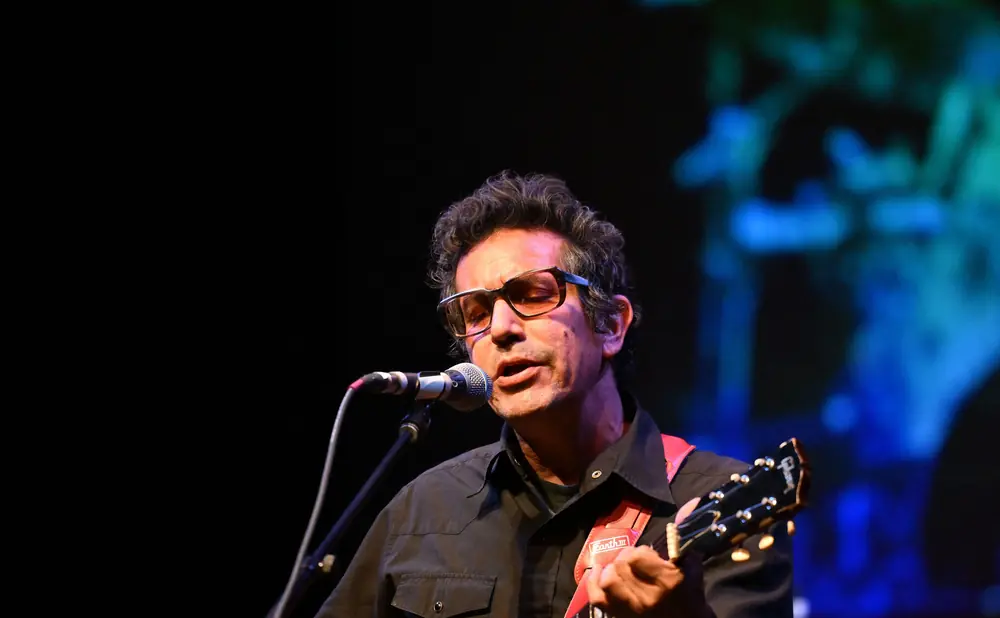 A man with glasses playing a red electric guitar and singing into a microphone on stage.
