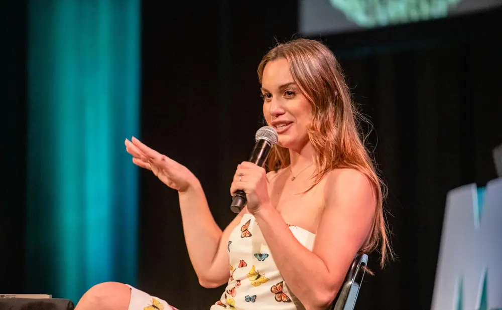 A woman in a strapless dress with butterflies on it holds a microphone and gestures with her hand.