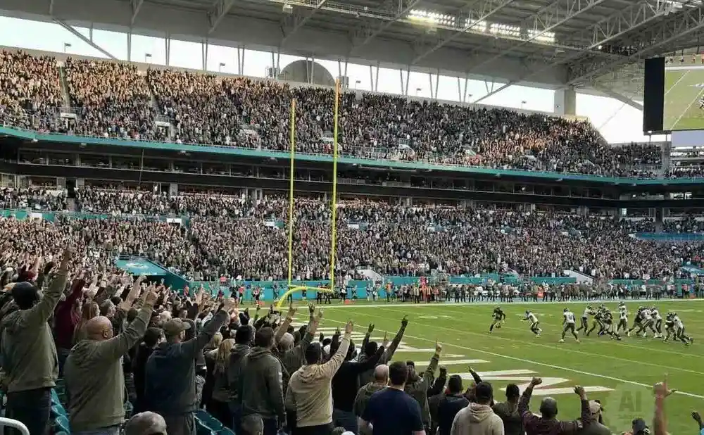 Fans cheer at a football game in a stadium, with players on the field and a large screen showing replays.