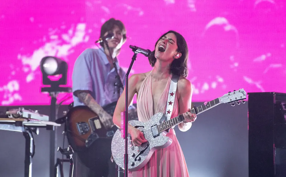 A female singer with a glittering guitar sings into a microphone while a male guitarist plays in the background.