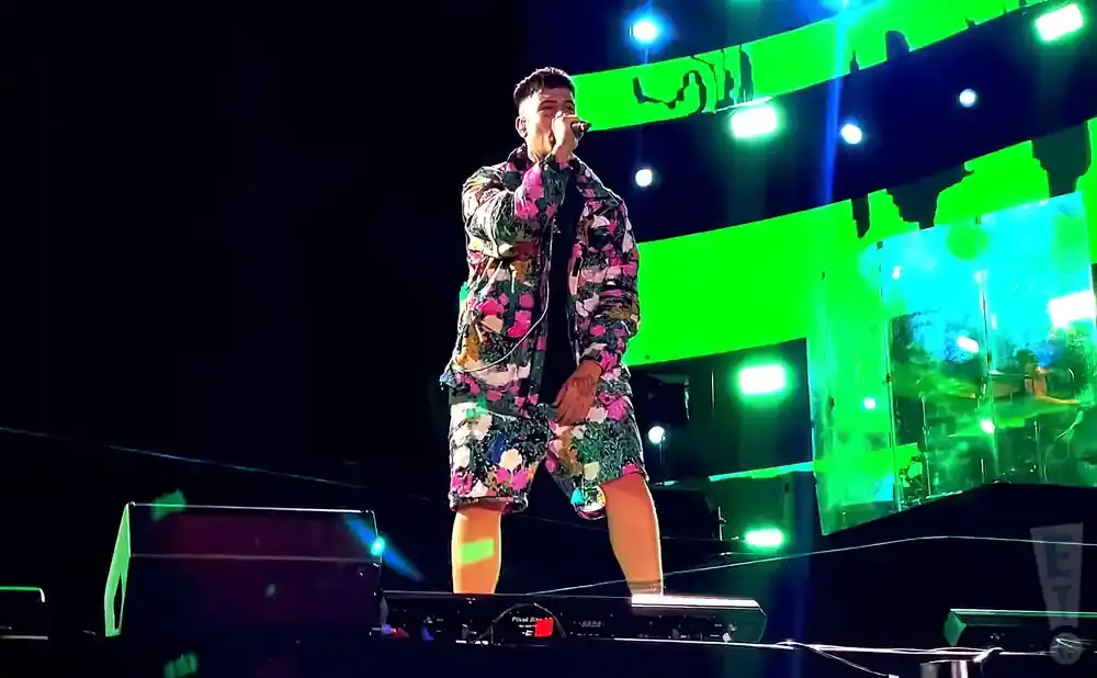 A male performer in a floral-print jacket and shorts sings into a microphone on a stage with a bright green backdrop.