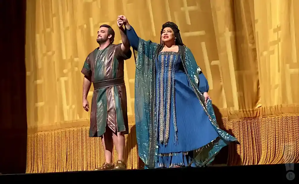 Two opera singers, a man in a bronze and green tunic and a woman in a blue gown, hold hands on stage.