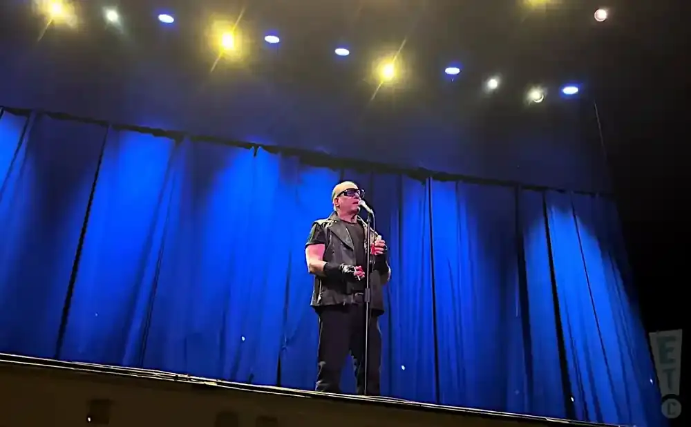 A bald man in sunglasses and a vest sings into a microphone on a stage with blue curtains.