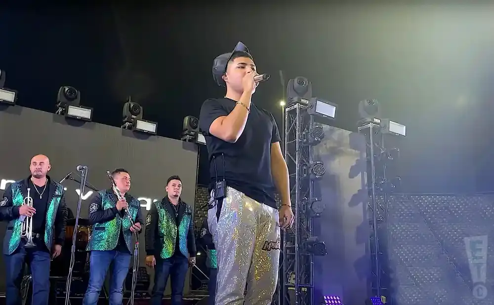 A singer in a black t-shirt and sparkly pants performs on stage with a band playing trumpets and a flute.