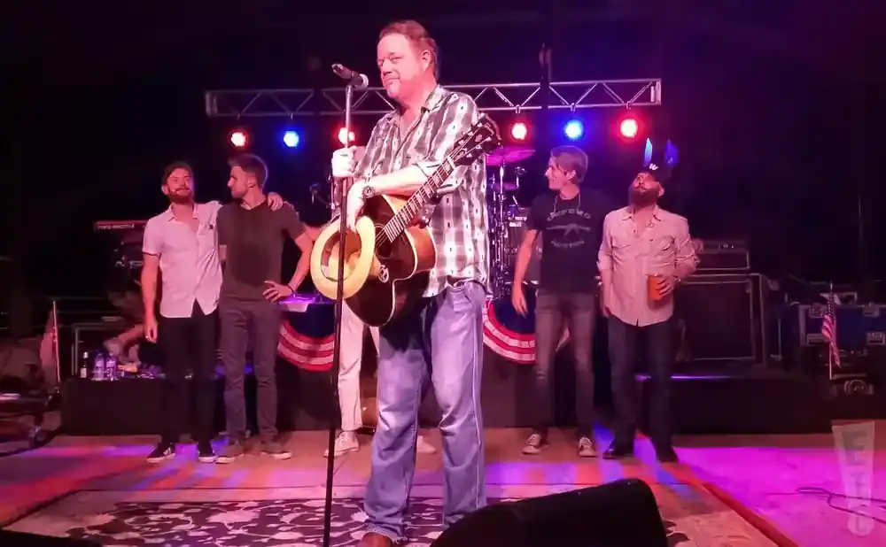 A man plays guitar and sings on stage while other band members stand around him.