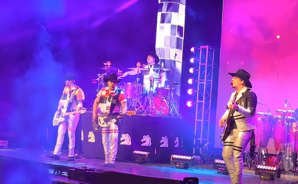 A band performs on a stage with purple and pink lights and fog. They wear matching sparkly outfits and cowboy hats.