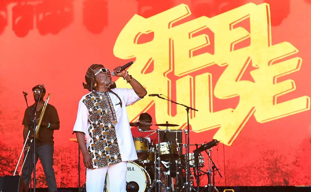 Reggae artist sings into a microphone on stage with a band, featuring a large yellow logo behind them.