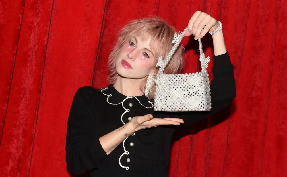 A woman with pink hair and star makeup holds a pearl beaded handbag in front of a red velvet curtain.